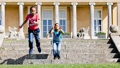 Children jumping down stone steps in front of a historic building with classical columns, outdoor activity, family fun, outdoor exploration, outdoor adventure.