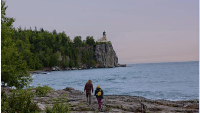 Seaside walk with lighthouse on rocky coast surrounded by lush greenery in outdoor adventure setting.