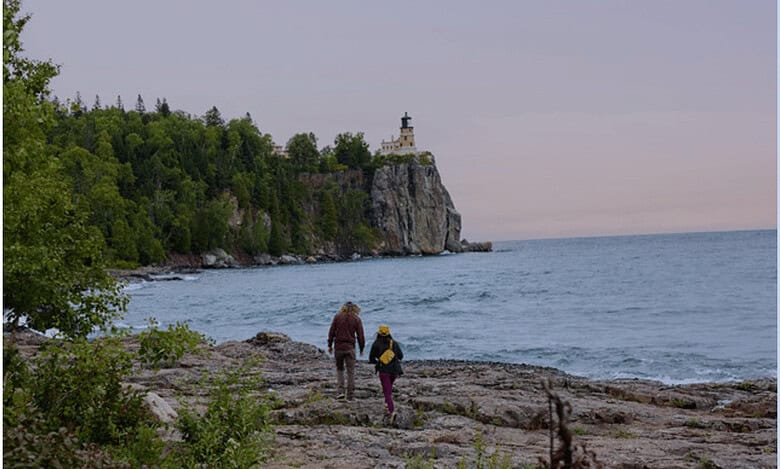 Seaside walk with lighthouse on rocky coast surrounded by lush greenery in outdoor adventure setting.