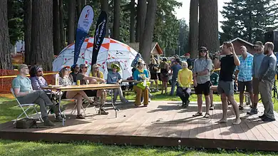 People listening to a speaker at an outdoor event in a forested area, with tents and banners in the background.