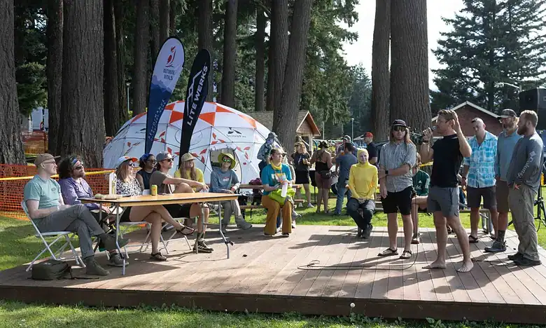 People listening to a speaker at an outdoor event in a forested area, with tents and banners in the background.