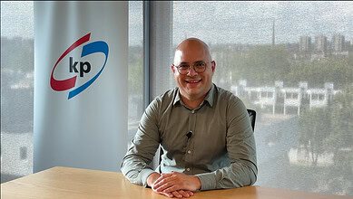 A man in glasses sitting at a desk with KP banner in the background, smiling in an office setting.