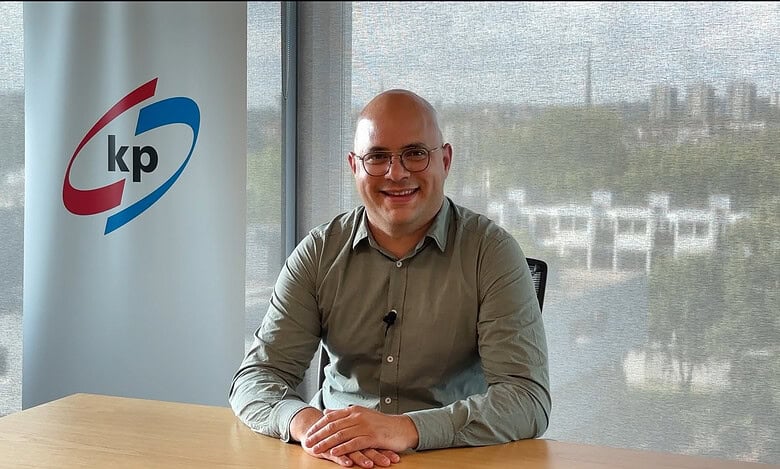 A man in glasses sitting at a desk with KP banner in the background, smiling in an office setting.