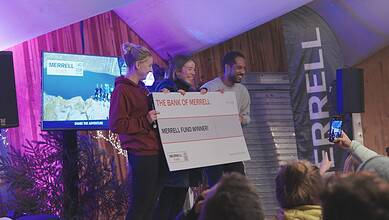 People celebrating outdoor adventure award win on stage at Merrell event in cosy wooden venue, smiling with large cheque.