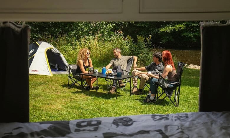Friends enjoying outdoor cooking and dining by a river with Campingaz grill and tent.