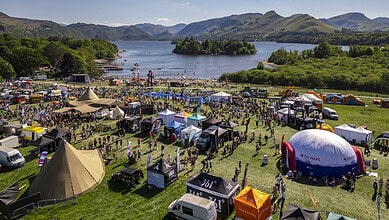 Outdoor festival with tents, stages, and crowds near a lake surrounded by mountains.