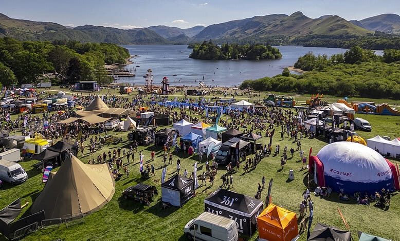 Outdoor festival with tents, stages, and crowds near a lake surrounded by mountains.