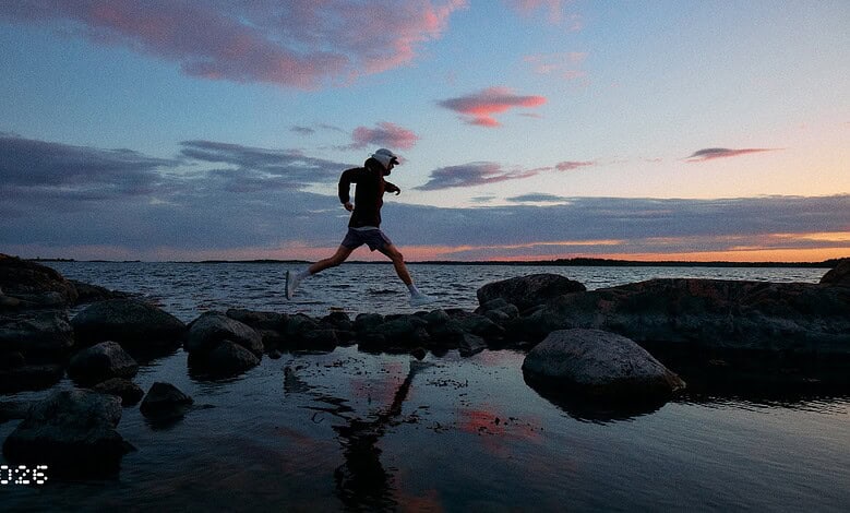 Person jogging on rocky beach during sunset with water and sky in background.