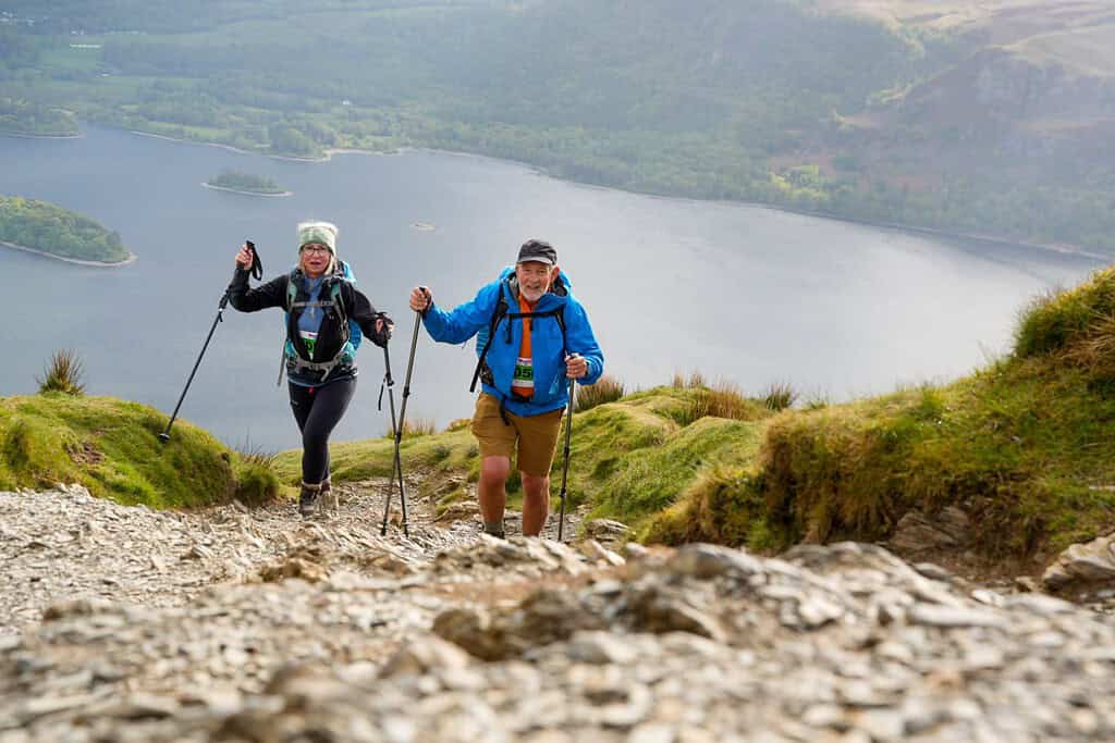 Climbers hiking on a rugged mountain trail with a lake view in the background.