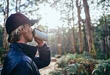 Person enjoying outdoor drink in forest during daytime, wearing outdoor gear.