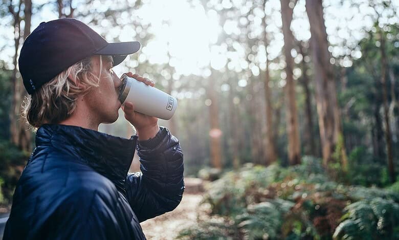 Person enjoying outdoor drink in forest during daytime, wearing outdoor gear.