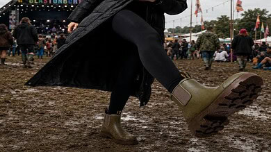 Woman in wellington boots at outdoor festival with muddy ground and stage in background.