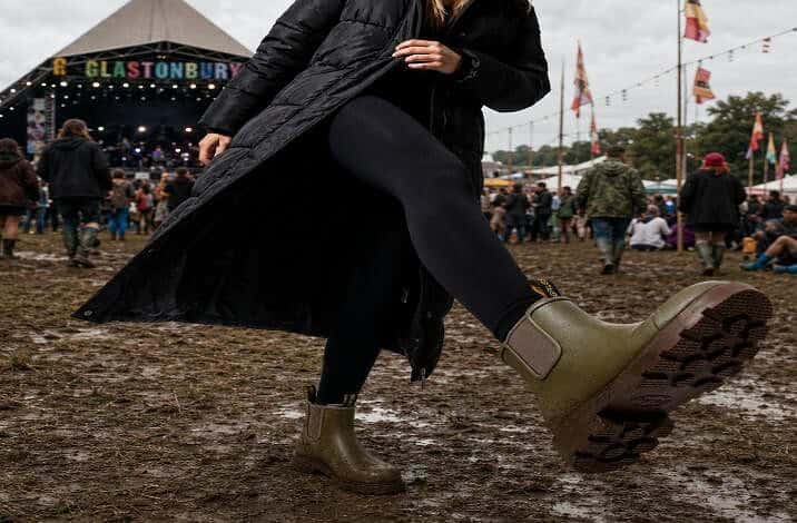 Woman in wellington boots at outdoor festival with muddy ground and stage in background.