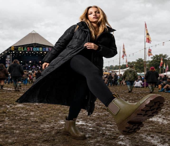 Woman in wellington boots at outdoor festival with muddy ground and stage in background.